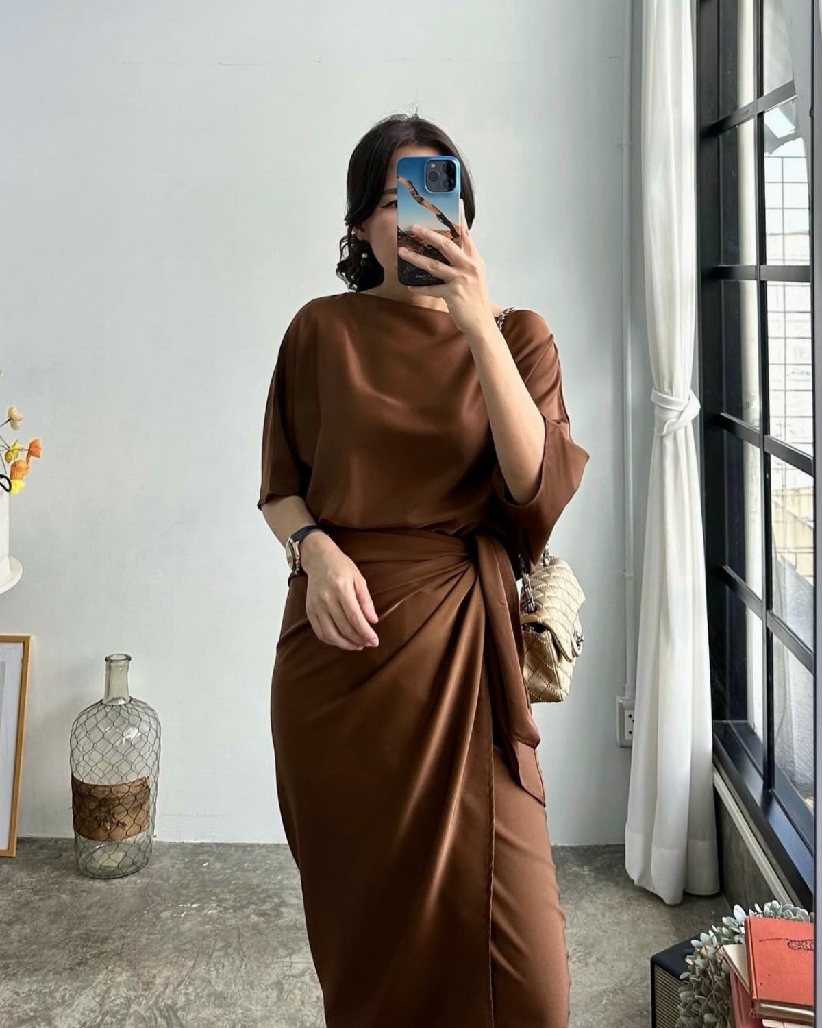 Woman in a brown satin wrap dress taking a mirror selfie indoors.