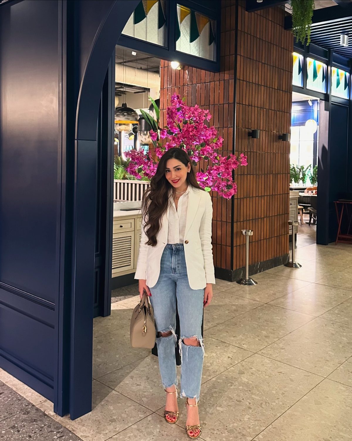 A woman posing in a white blazer, white top, and ripped blue jeans with a tan handbag.