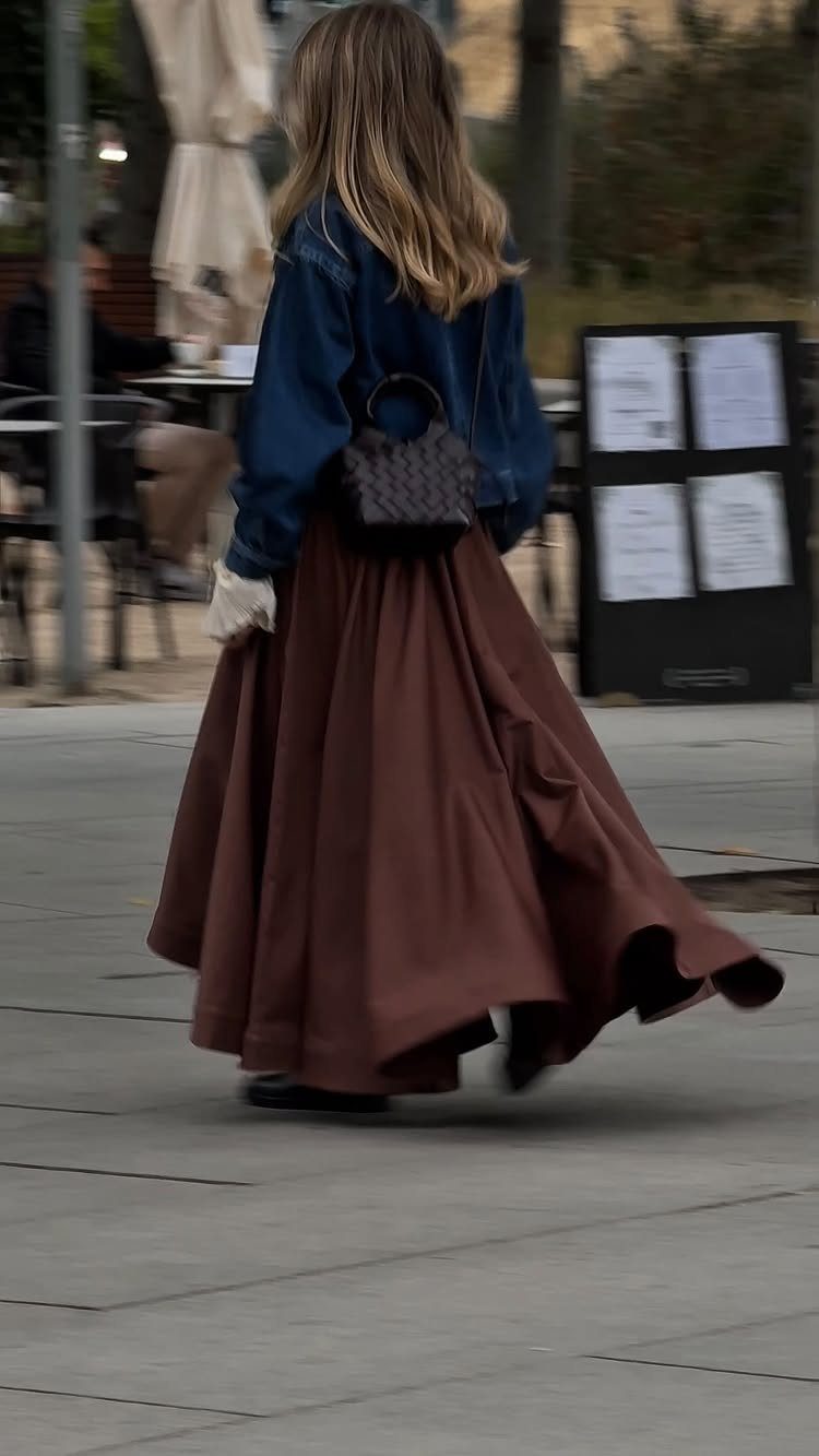 A woman walking in a denim jacket and a long brown flowing skirt.