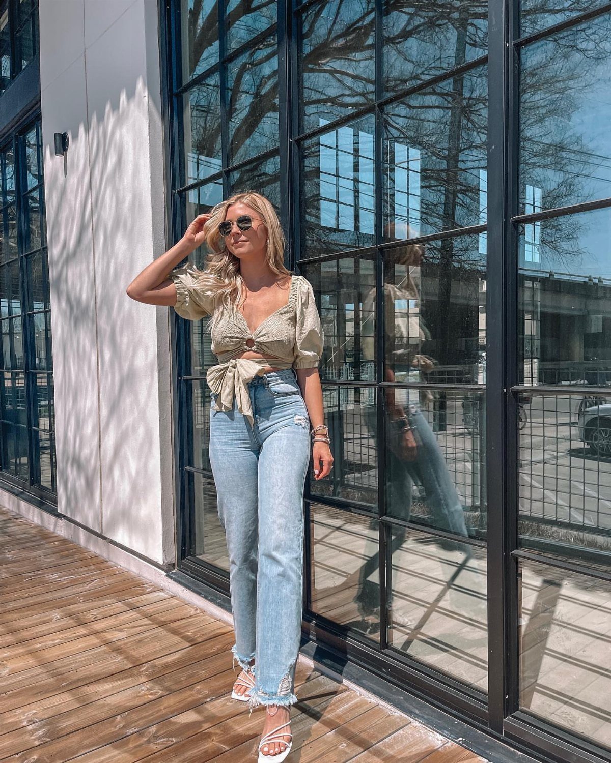 A blonde woman posing in a green tie-front top and light blue jeans outdoors.