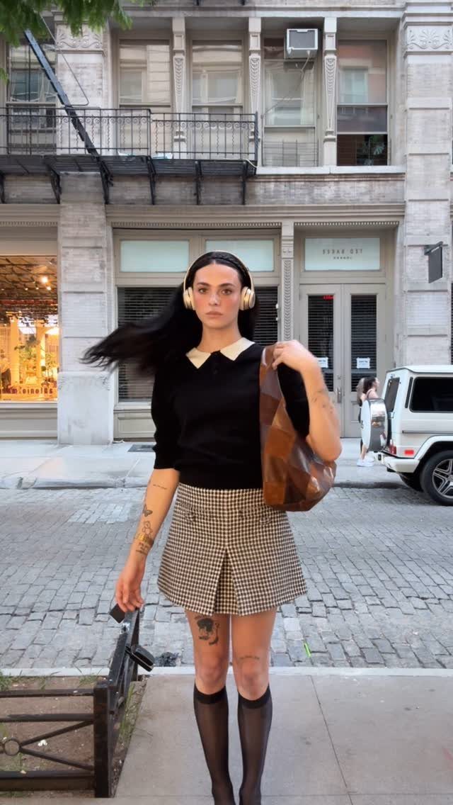 Woman in a black collared top, plaid skirt, and knee-high socks standing on a city street.