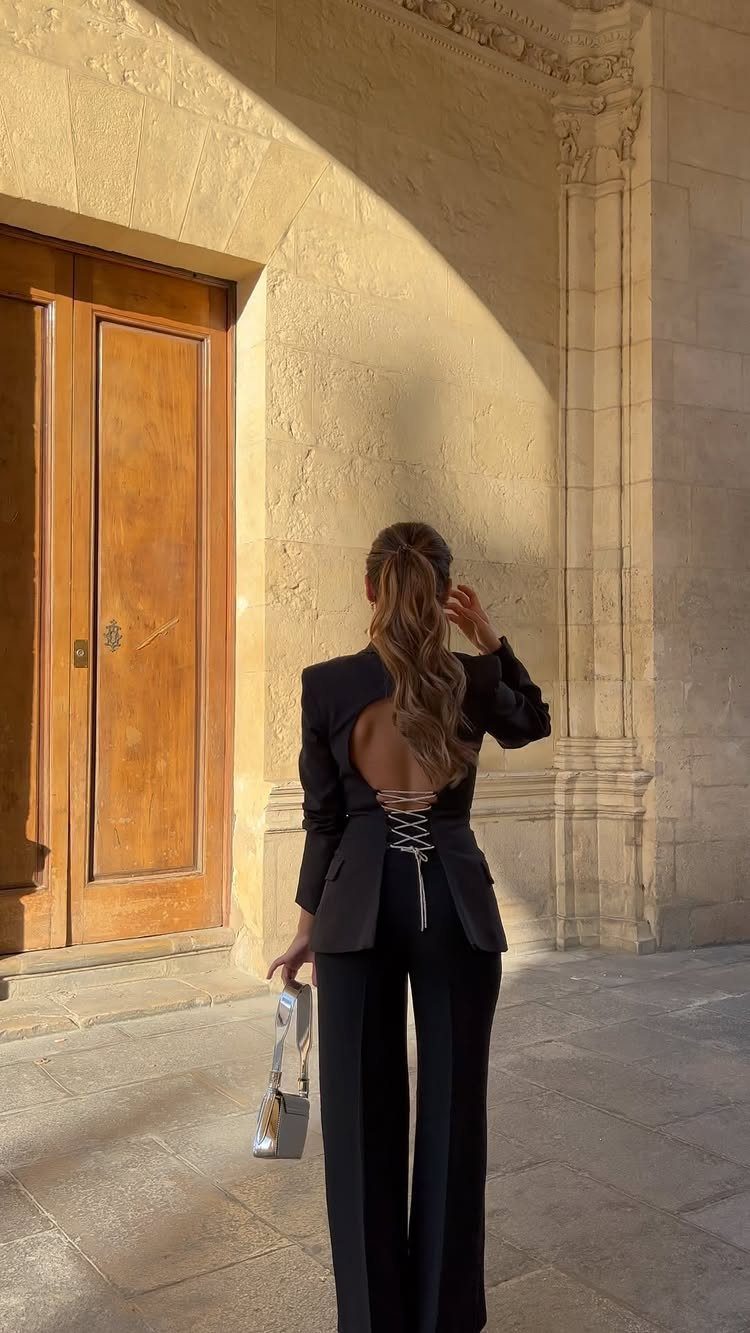 Woman in a black lace-up back blazer and trousers standing by a stone wall.