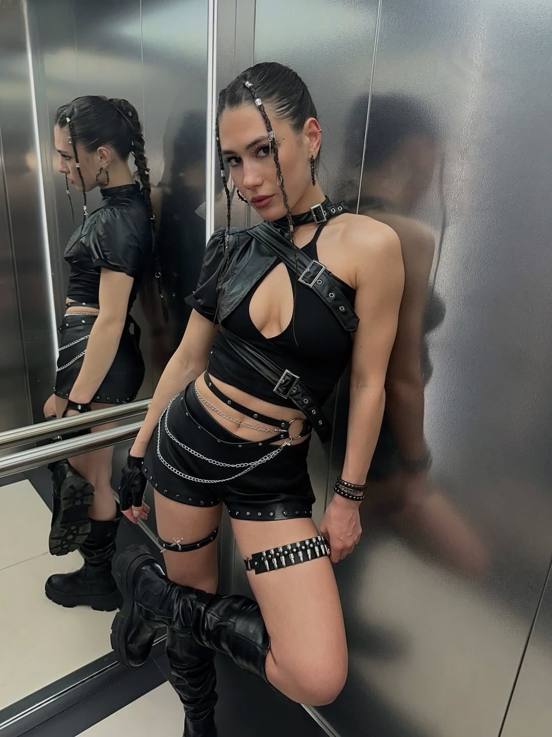 Woman in a black leather rave outfit posing in a metallic elevator with silver chain details.