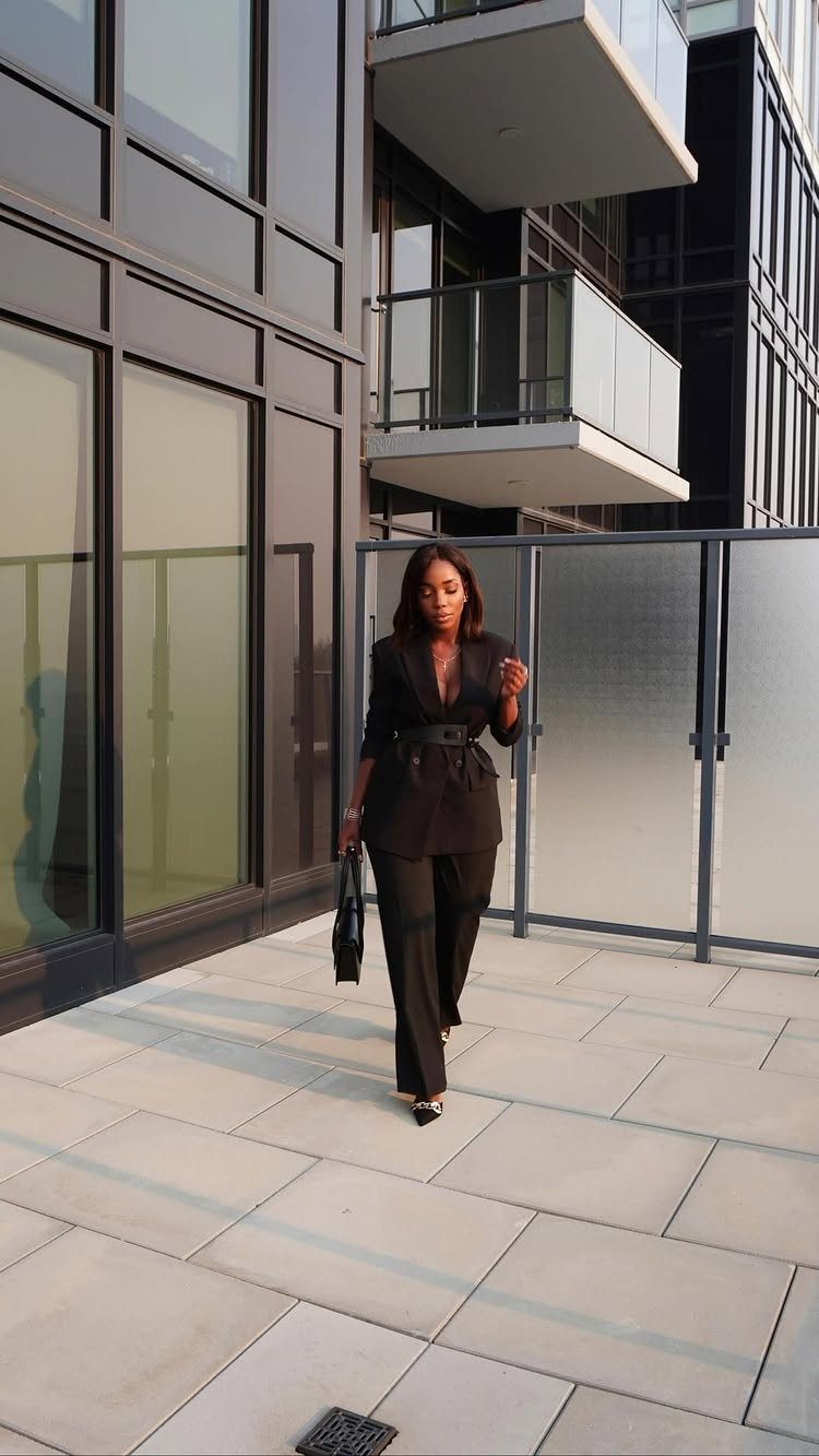 A woman in a black belted blazer and matching trousers walking on a modern terrace.