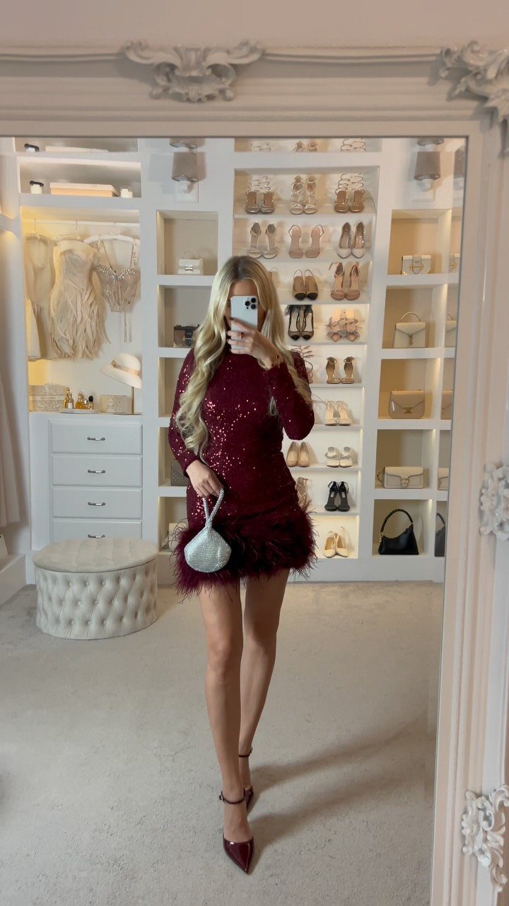Woman in a sequined maroon mini dress with feather trim posing in a walk-in closet.