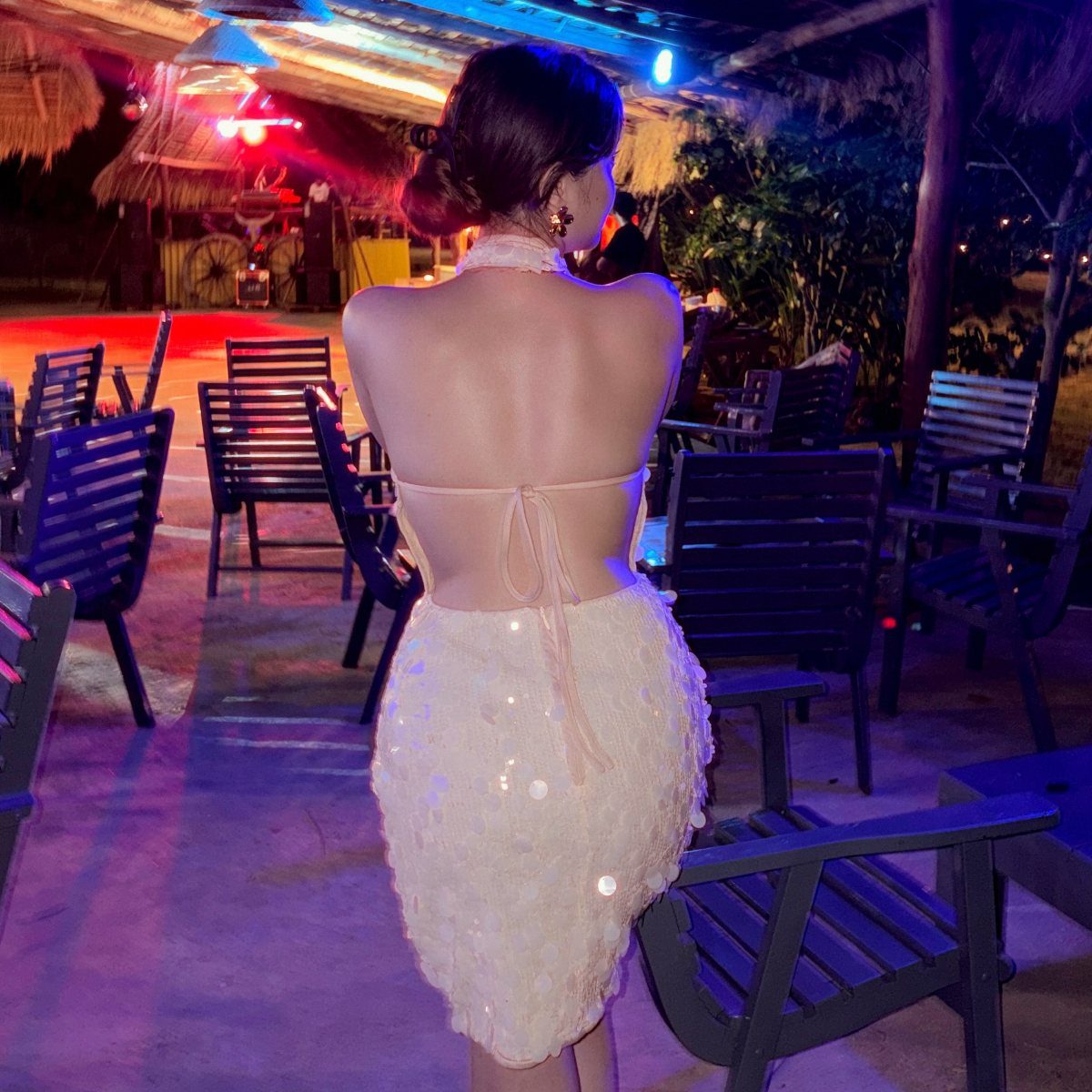 Woman in a white sequined halter-neck mini dress standing in an outdoor patio at night.