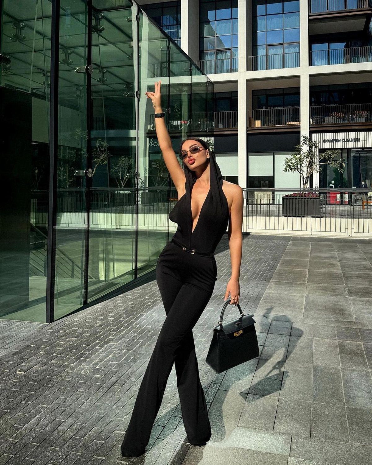 Woman in a black sleeveless jumpsuit posing outdoors in front of a glass building.