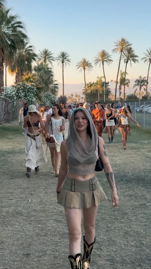 A woman in a hooded crop top and tan skirt walking through a palm-lined festival field.
