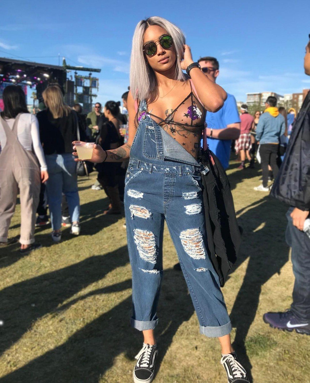 Woman in distressed denim overalls and black sneakers standing in a sunny outdoor festival field.