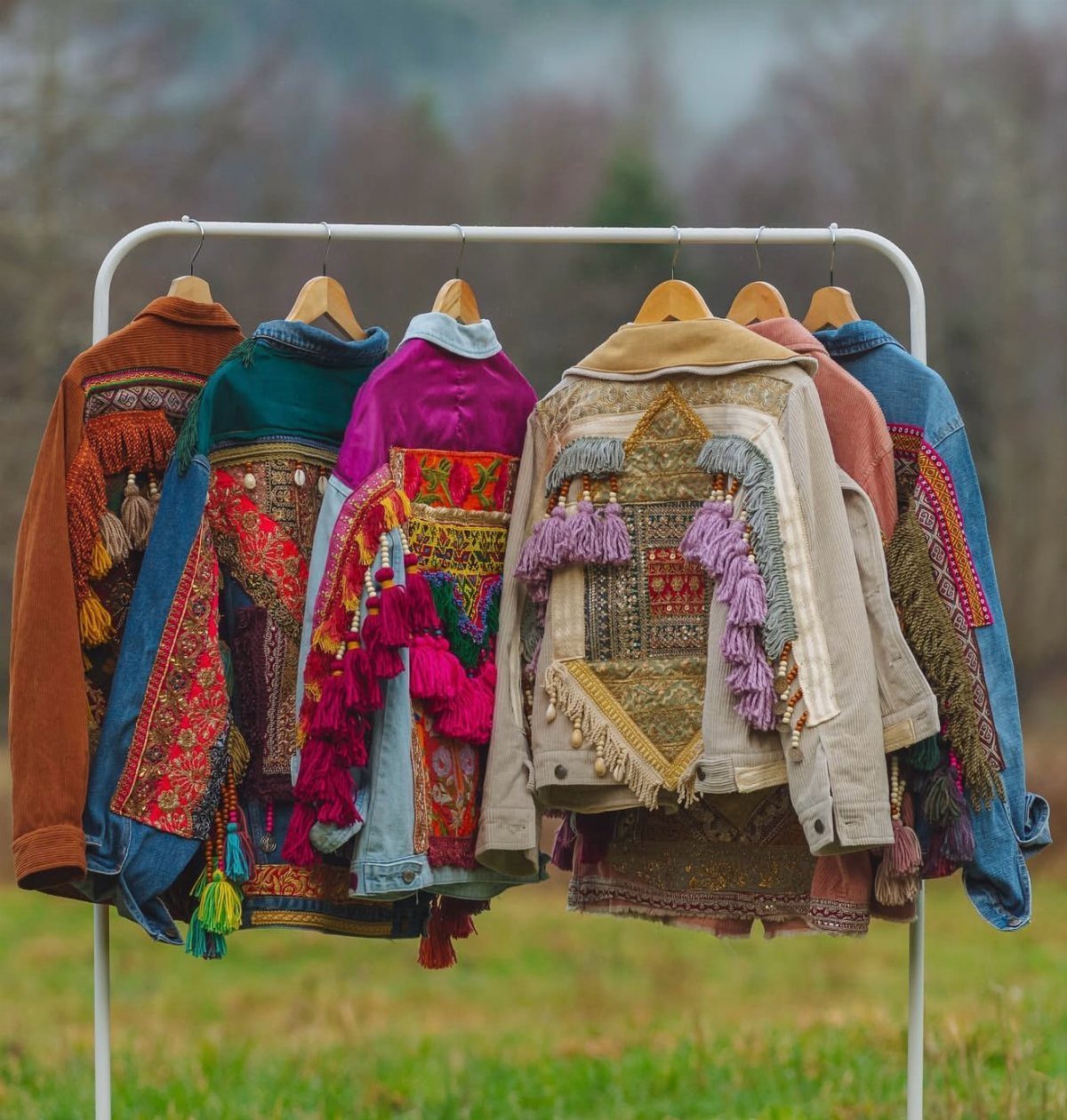 Six colorful bohemian embroidered jackets hanging on a white clothing rack in a grassy field.