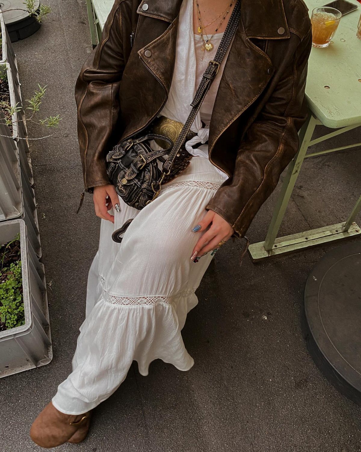 A person wearing a white flowy maxi dress and a brown leather jacket sitting outdoors.