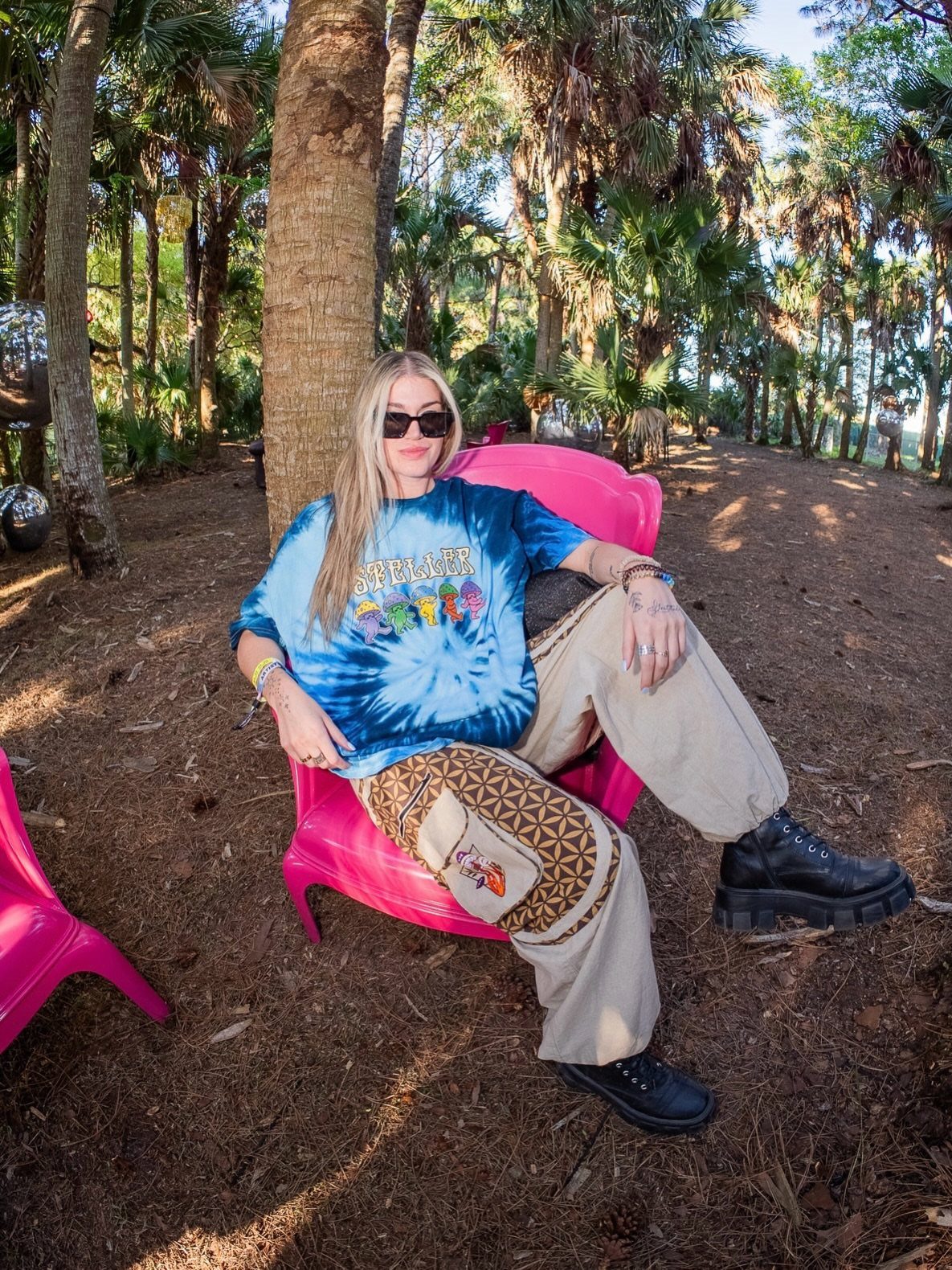 A woman wearing a blue tie-dye graphic band tee and patterned cargo pants sitting in a pink chair.