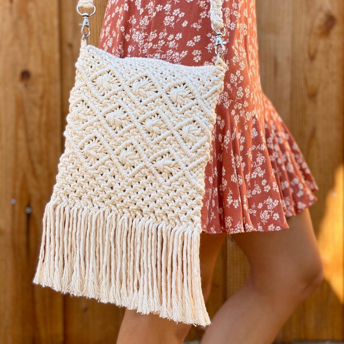 A cream macrame fringe bag worn over a floral patterned dress against a wooden background.