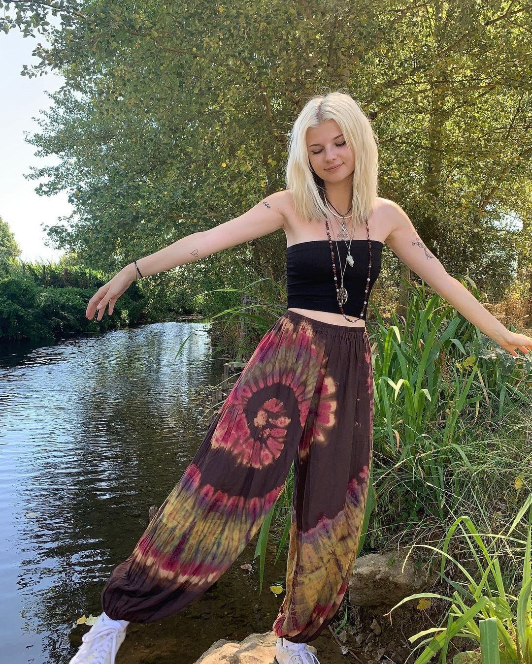 Blonde woman in tie-dye pants and black tube top standing by a river.