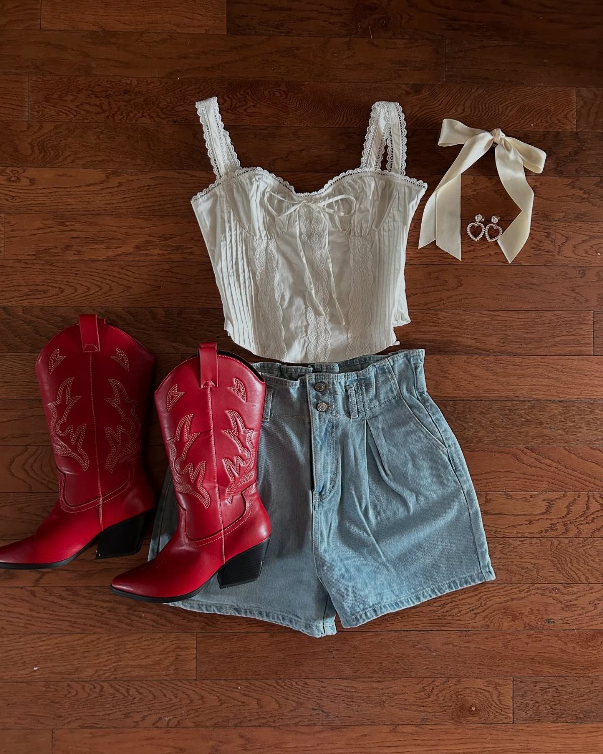 White lace camisole, denim shorts, red cowboy boots, and accessories laid on a wooden floor.