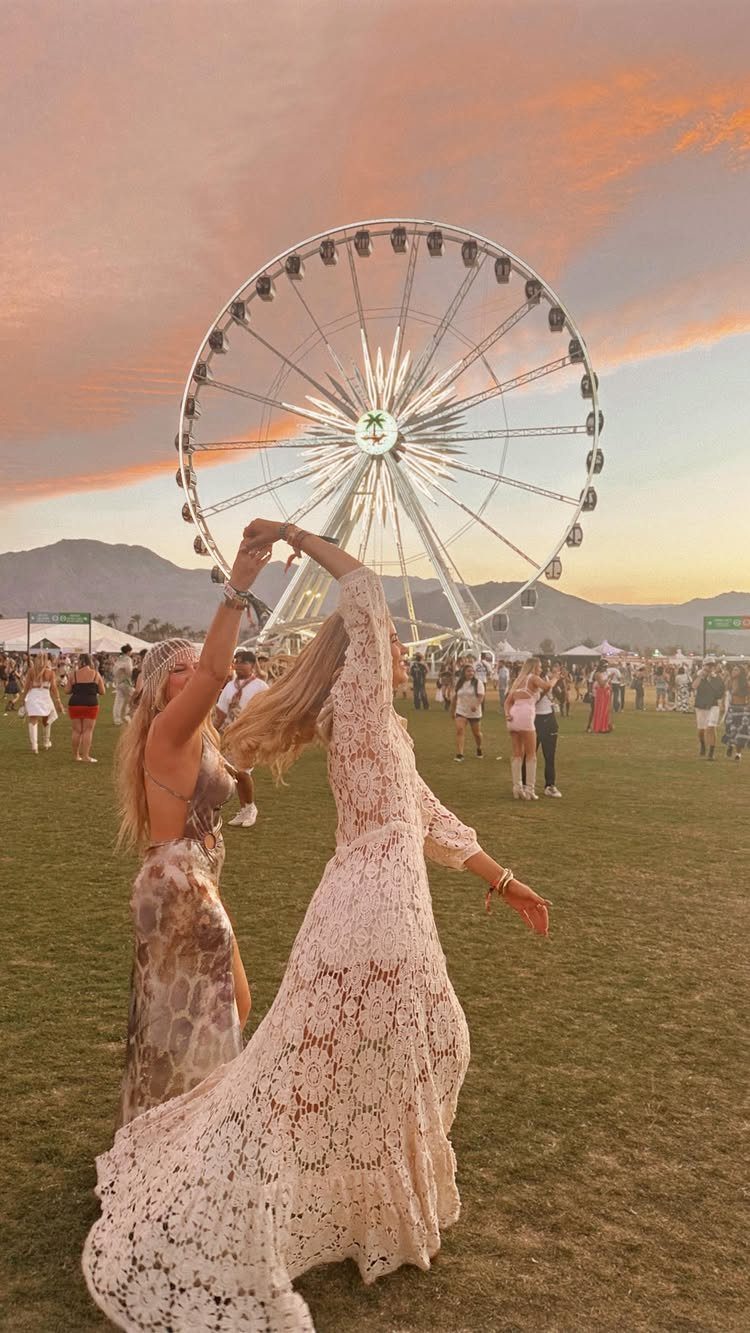 Two women dancing in a field with a large Ferris wheel at sunset.