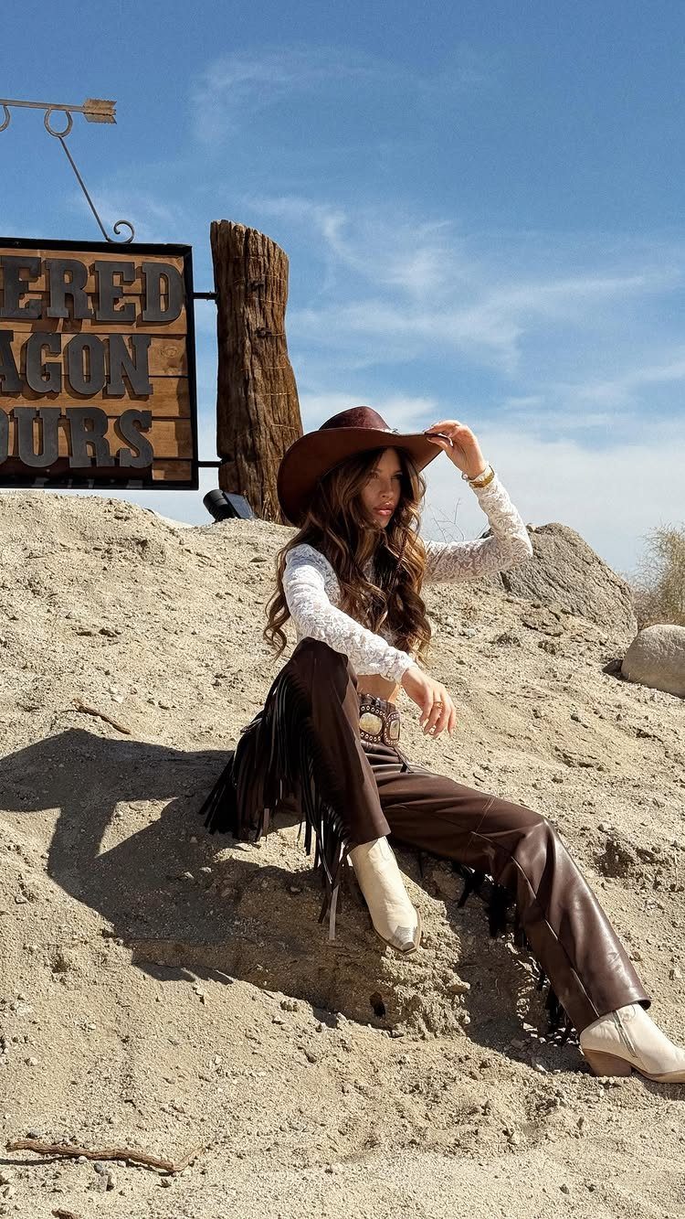 Woman in western attire sitting on a sandy hill wearing a cowboy hat and fringe pants.
