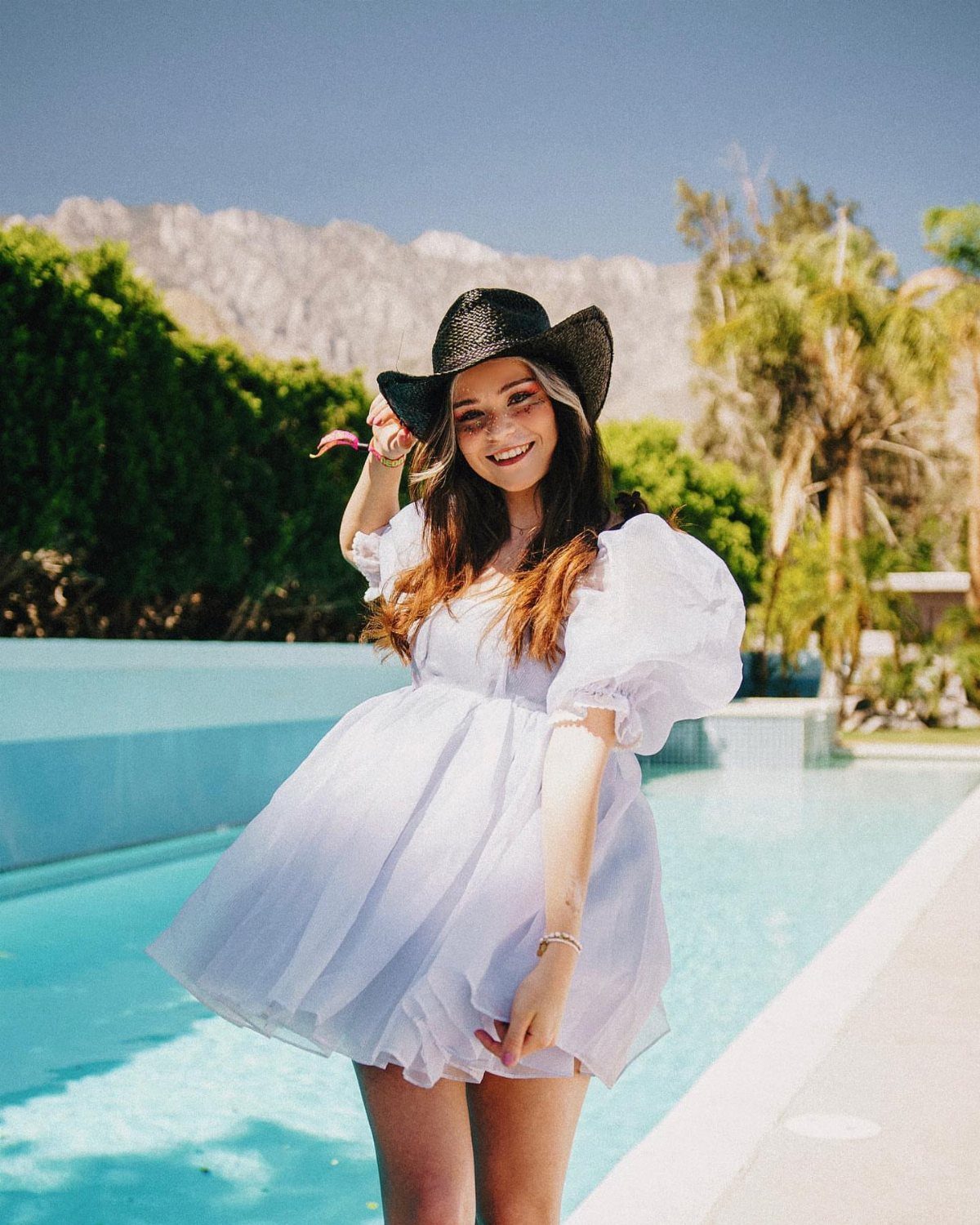 A woman in a white puff-sleeve dress and black cowboy hat posing by a pool.