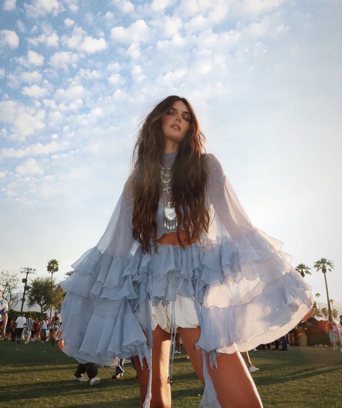 Woman in a ruffled light blue tiered outfit posing under a cloudy blue sky.