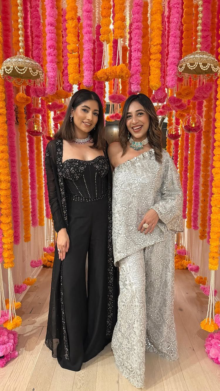 Two women posing in elegant black and silver sequined jumpsuits against a floral backdrop.