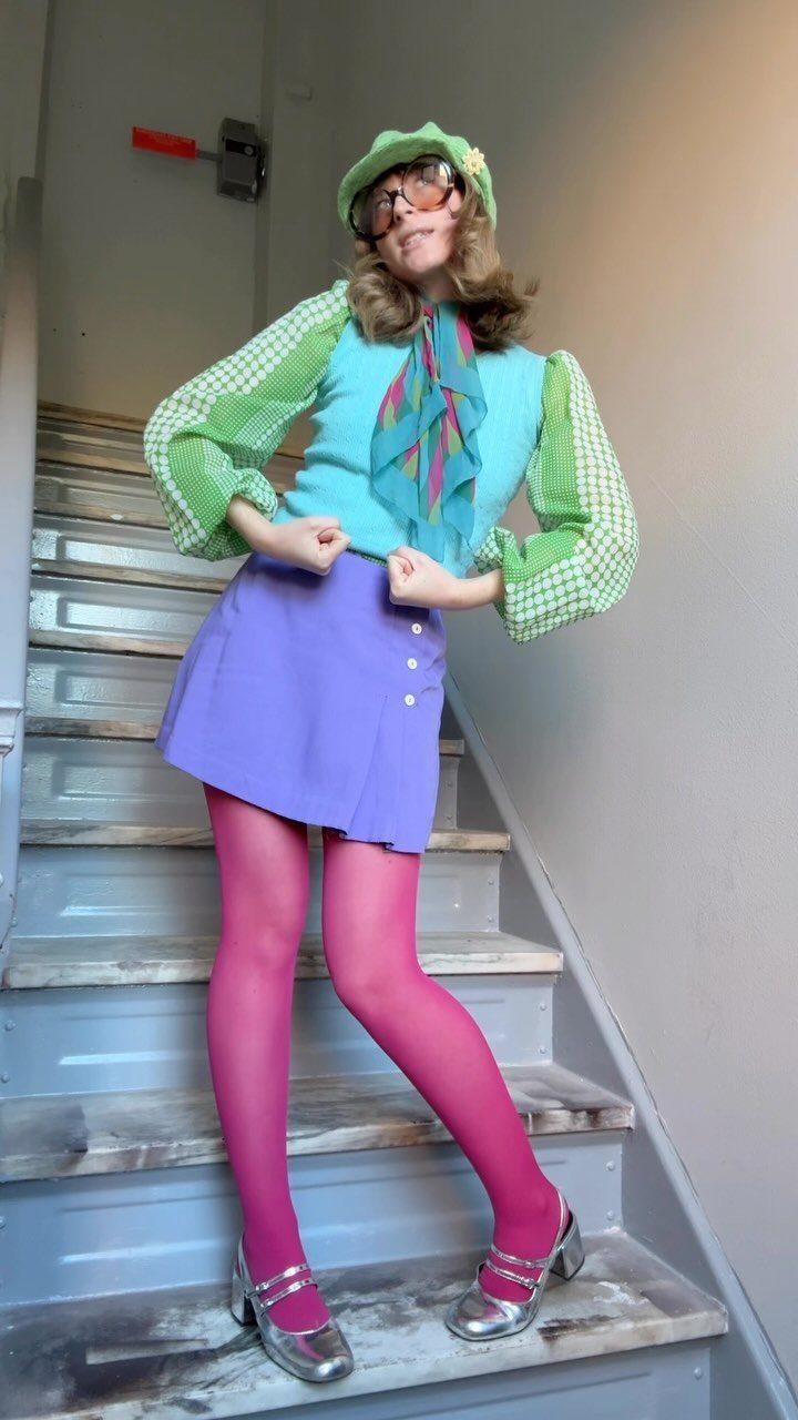 Woman in colorful turquoise vest, purple skirt, and pink tights posing on a staircase.