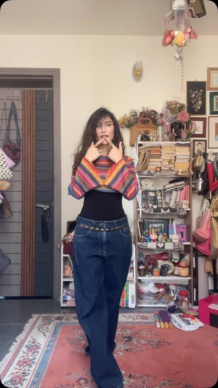 A woman posing in a colorful crochet shrug, black top, and blue denim jeans.
