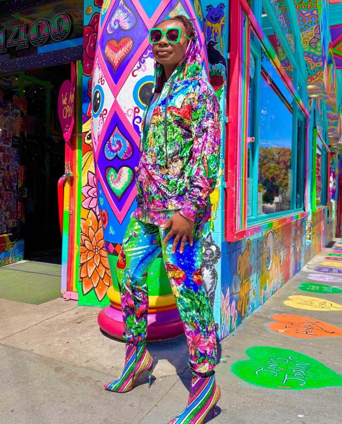 A person in a colorful patterned tracksuit and green sunglasses posing against a psychedelic mural.