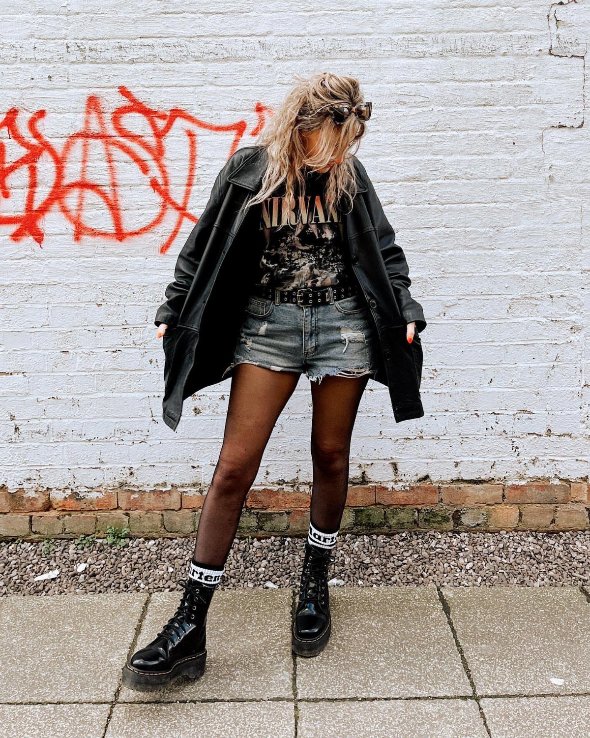 A person wearing a graphic tee, denim shorts, black tights, and black combat boots against a graffiti wall.