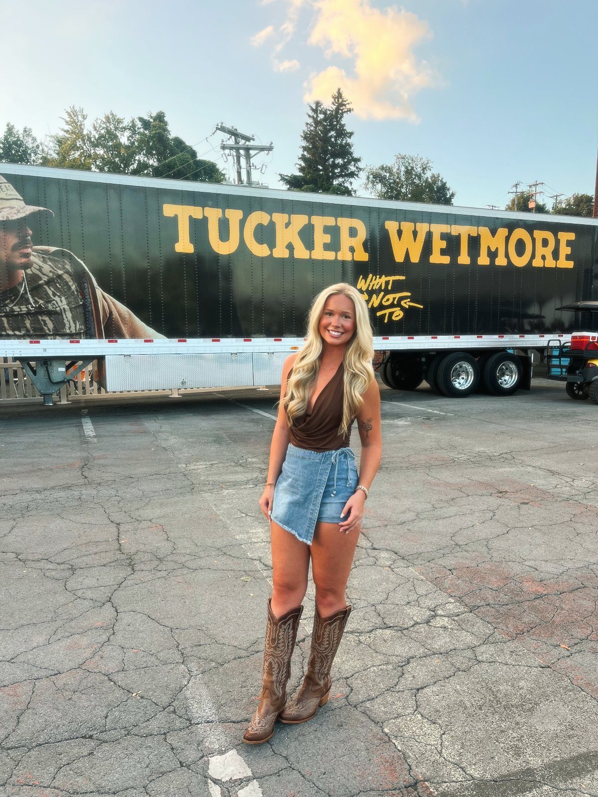 Blonde woman in a denim skirt and patterned knee-high cowboy boots standing in a parking lot.