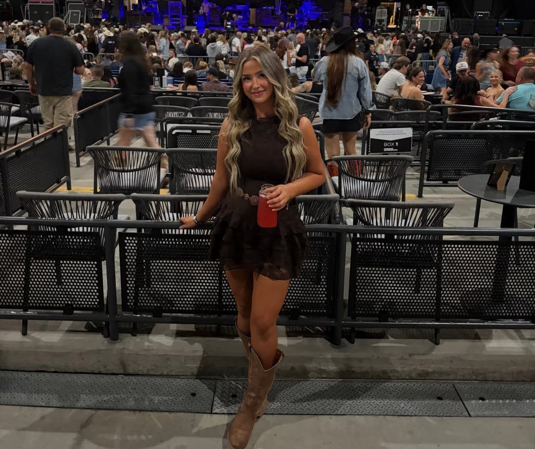 Woman in a dark brown mini dress and tan cowboy boots posing at a concert.