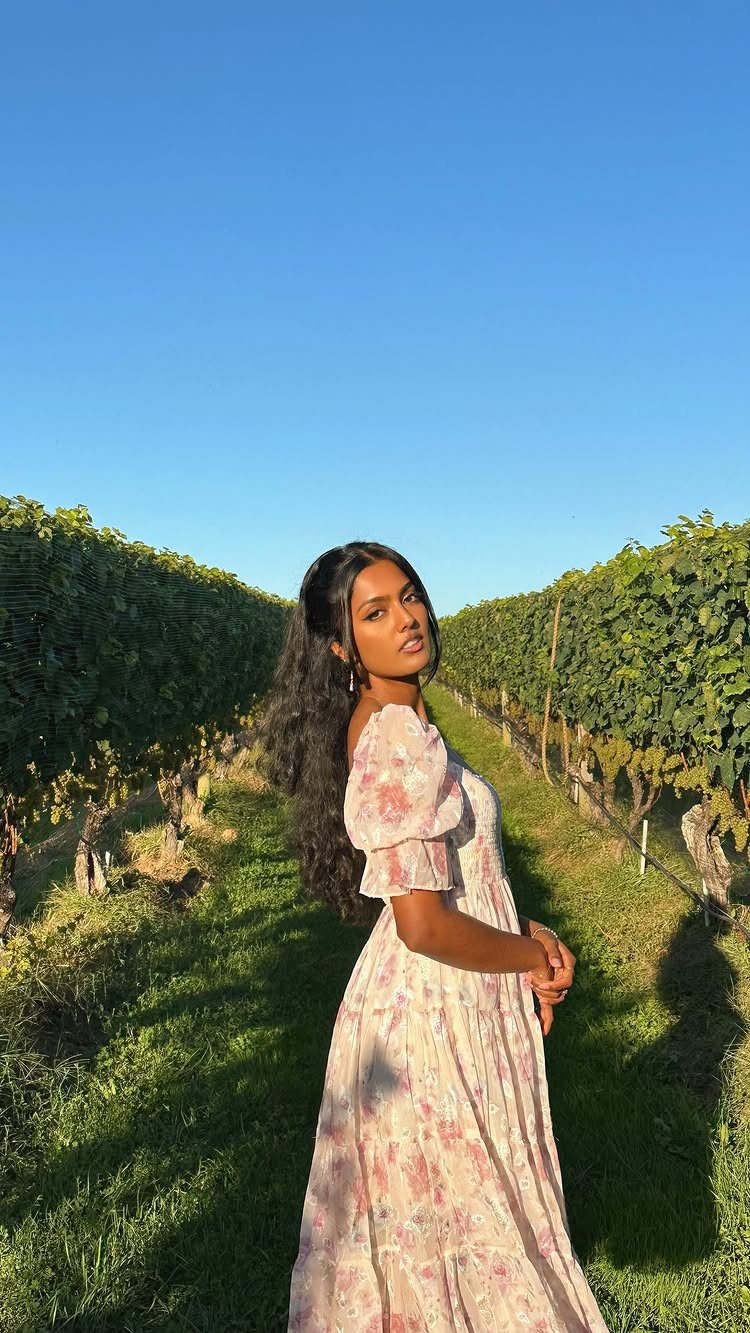 A woman in a floral flowy dress posing in a sunny vineyard between rows of vines.
