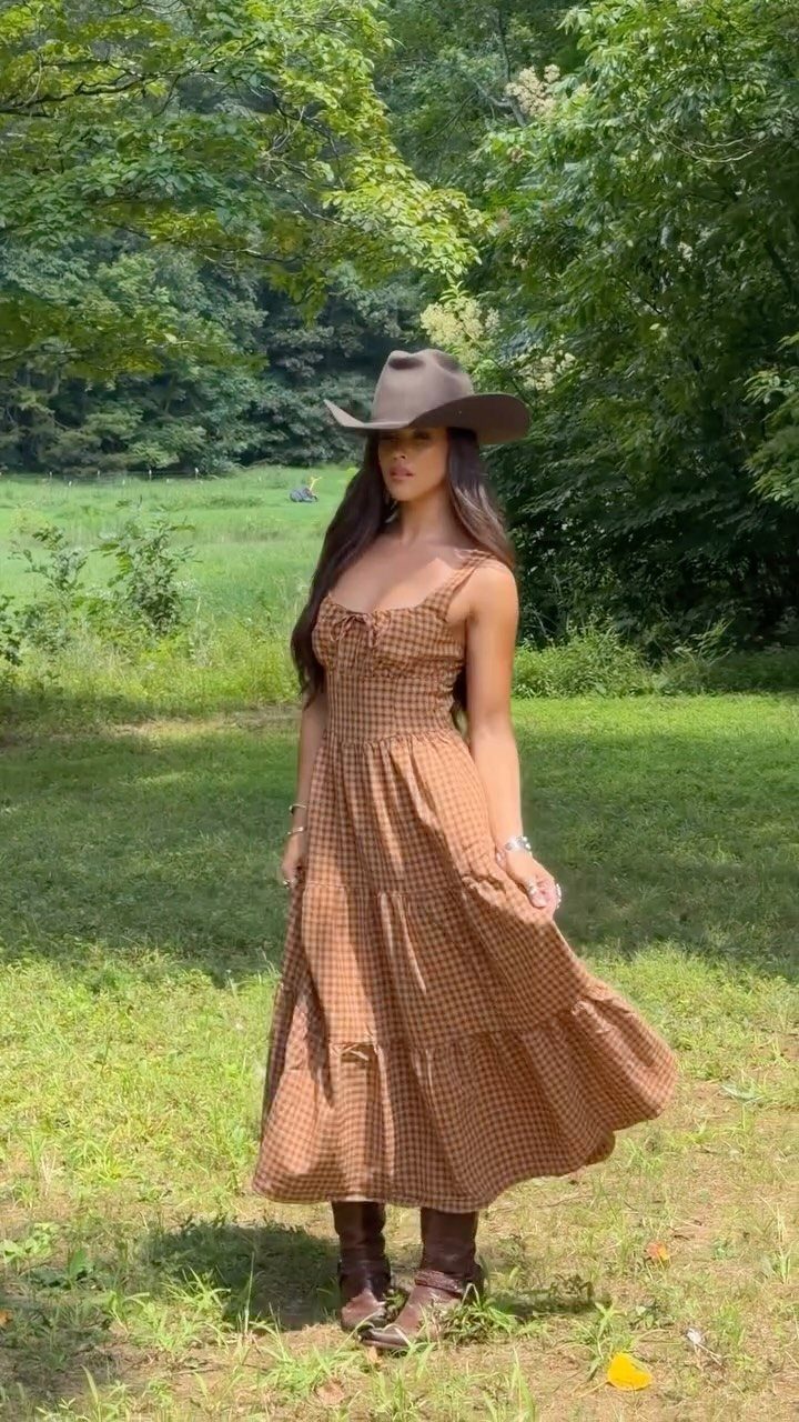 A woman in a brown patterned maxi dress and cowboy hat standing in a grassy field.