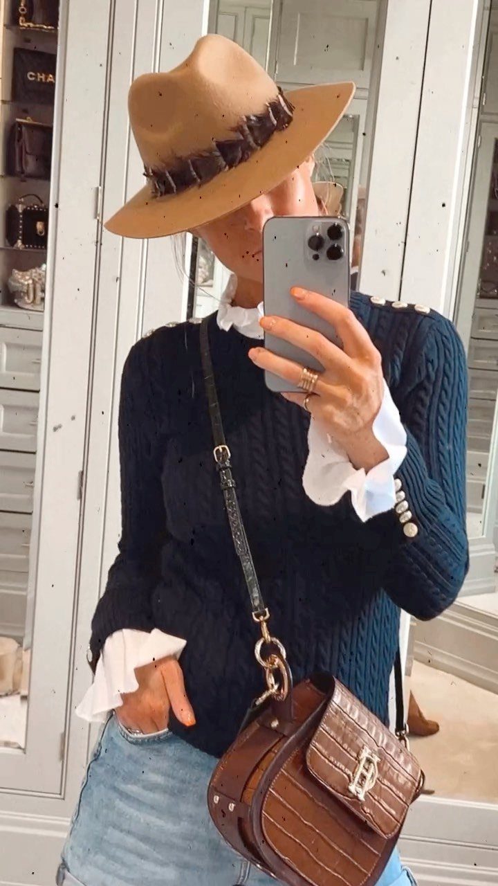 Person in a blue sweater, tan hat, and denim shorts taking a mirror selfie.