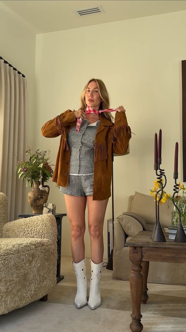 Woman posing in a brown fringed jacket, patterned top, denim shorts, and white boots.