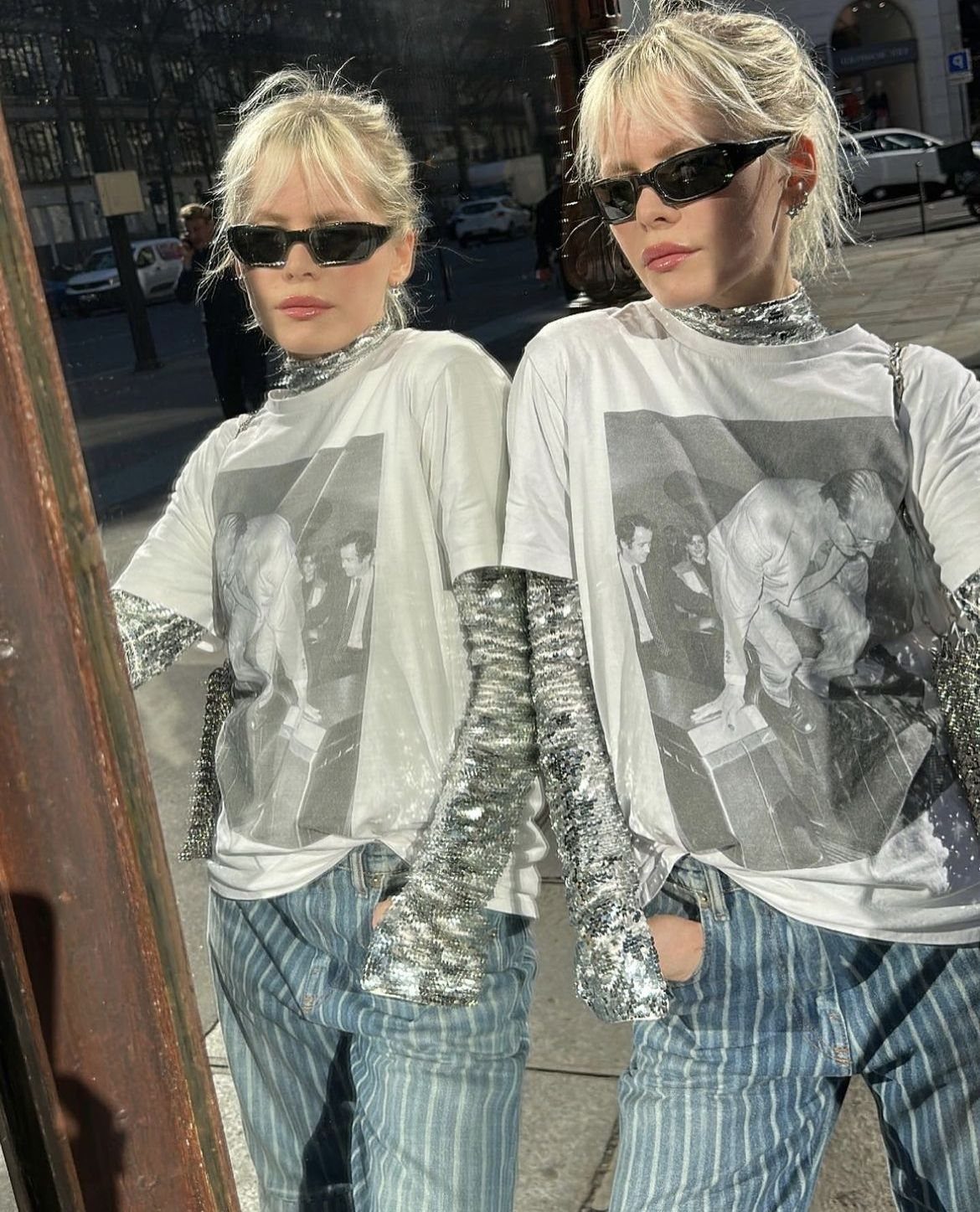 Person in a graphic tee and silver sequined sleeves posing in a mirror outdoors.