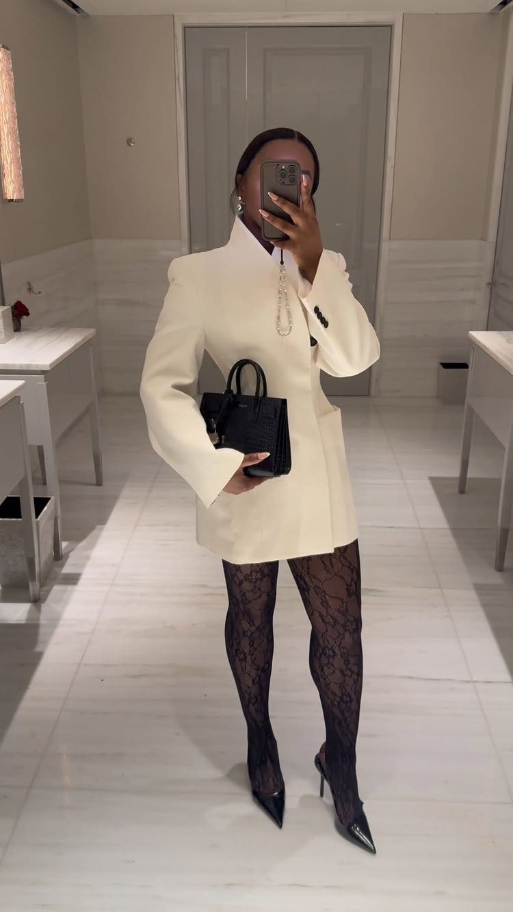 A person in a cream oversized blazer, lace tights, and black heels holding a handbag.