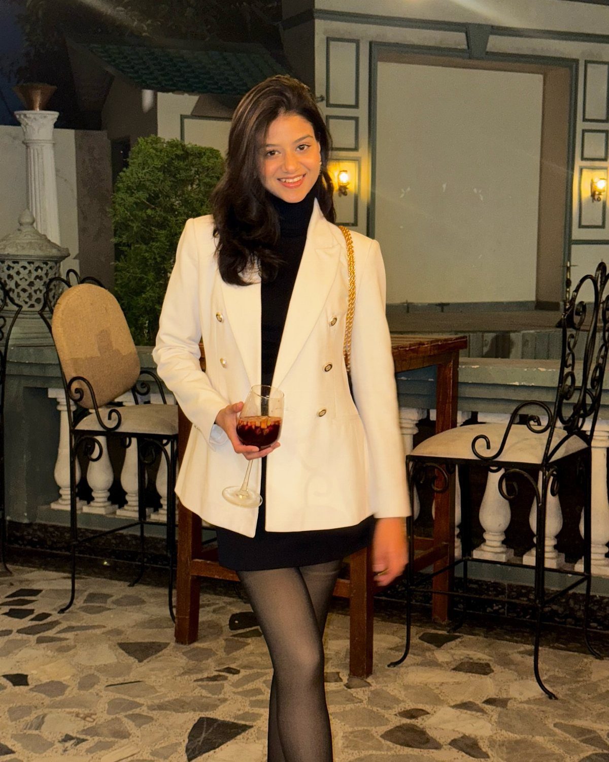 A smiling woman wearing a white oversized blazer over a black dress and black tights.