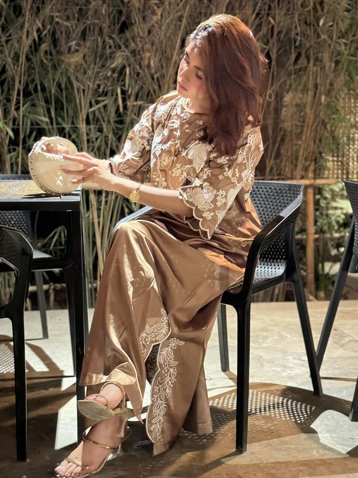 A woman in a bronze two-piece lace outfit sits at an outdoor cafe table.
