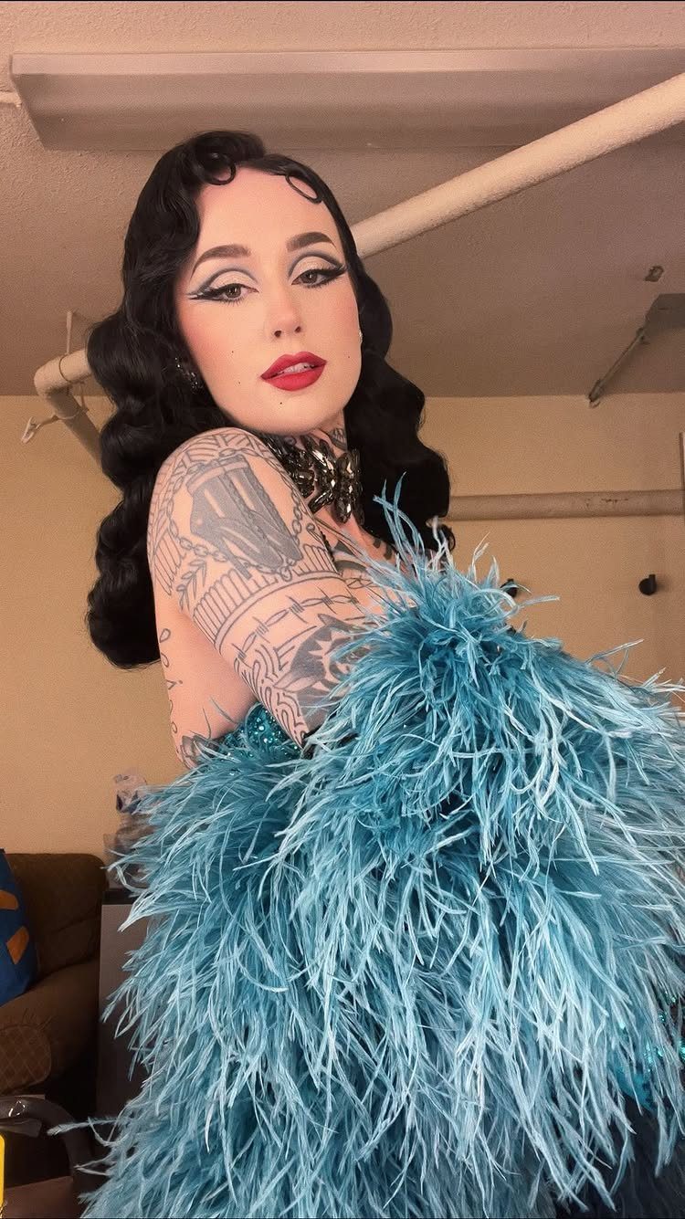 A woman with dark hair and tattoos wearing a large teal feathered outfit.