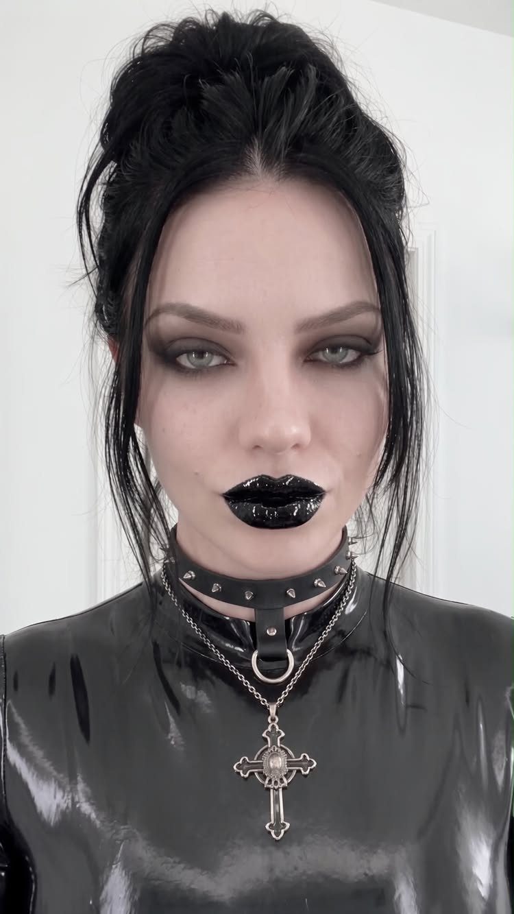 Person with dark hair and heavy makeup wearing a black latex top and silver cross necklace.