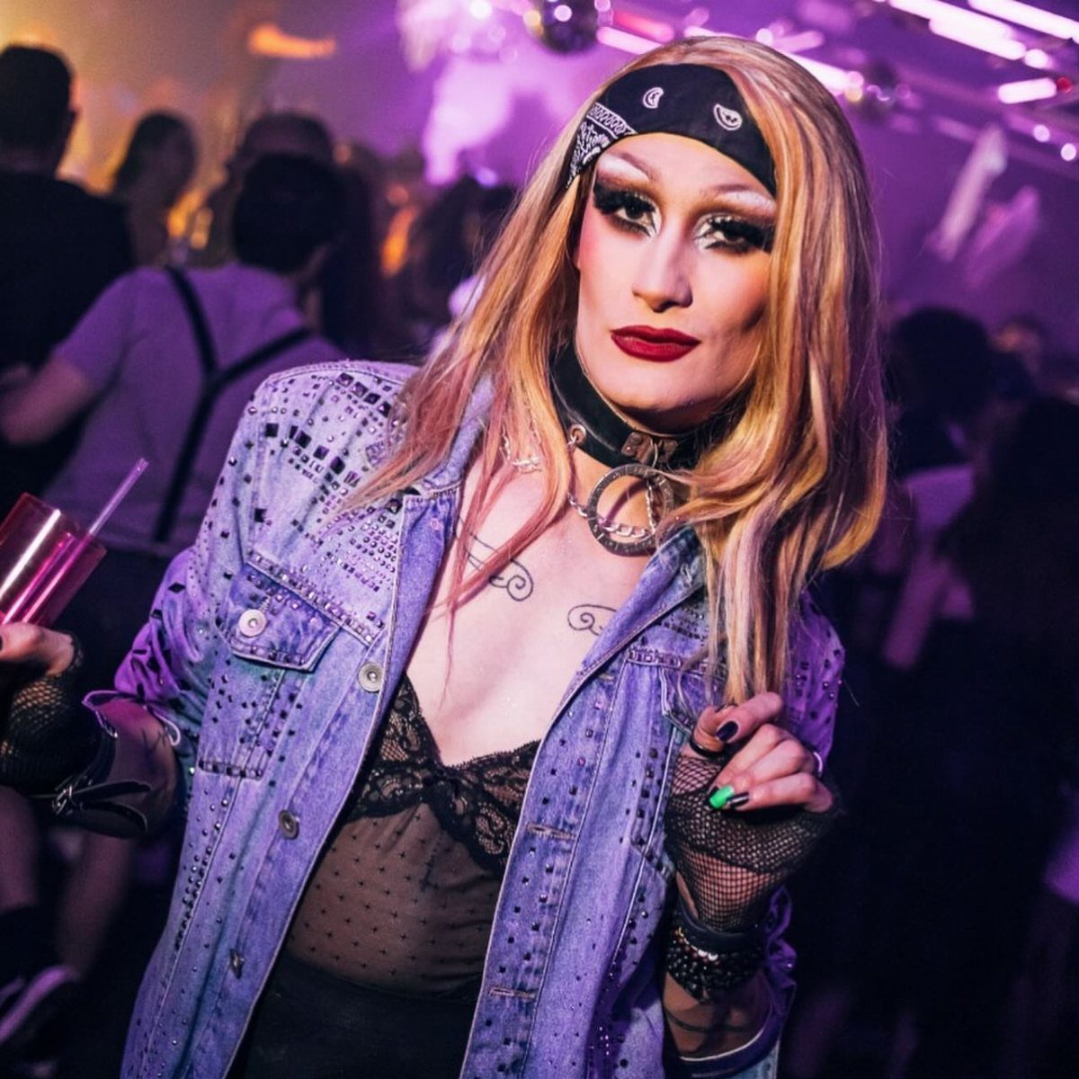 A person with dramatic makeup and a studded denim jacket posing in a nightclub setting.