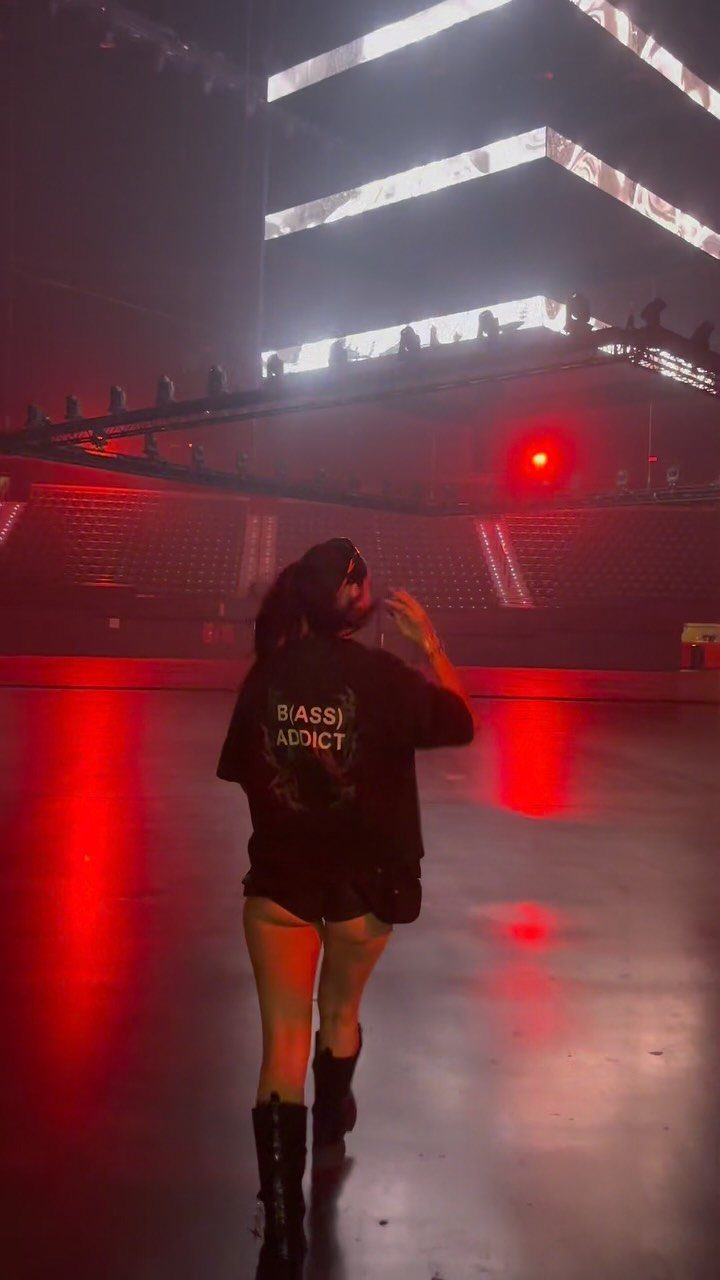 A person wearing a black graphic t-shirt and shorts stands in a large, red-lit concert venue.