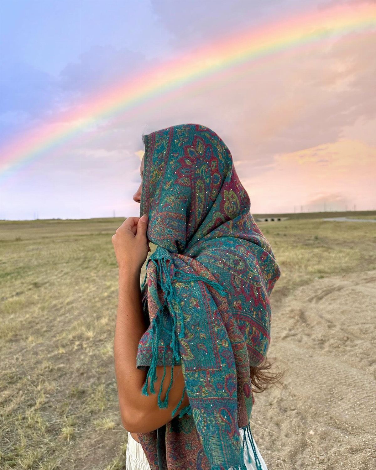 Person wearing a patterned teal and red pashmina under a large rainbow in a field.