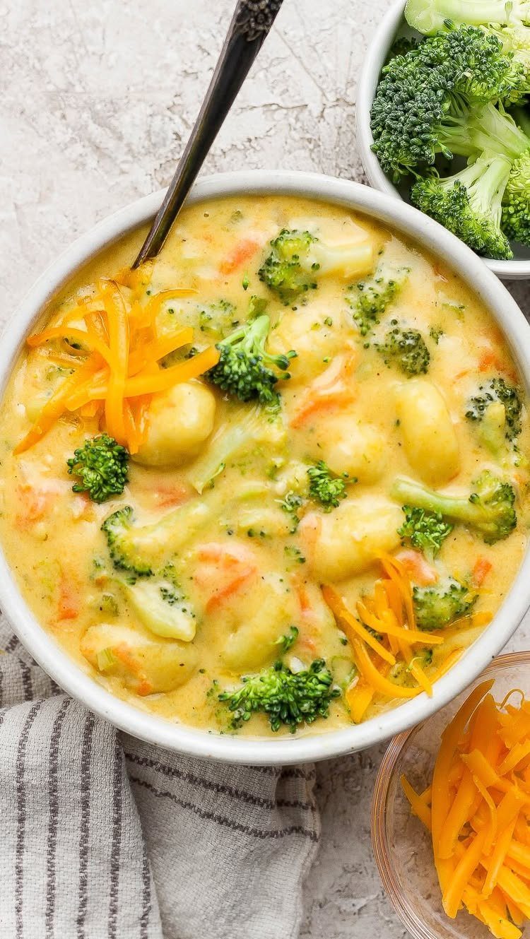 Creamy broccoli cheddar gnocchi soup in a white bowl with shredded cheese and broccoli florets.