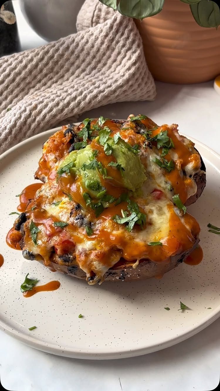 Stuffed sweet potato topped with melted cheese, red sauce, guacamole, and fresh cilantro on a plate.