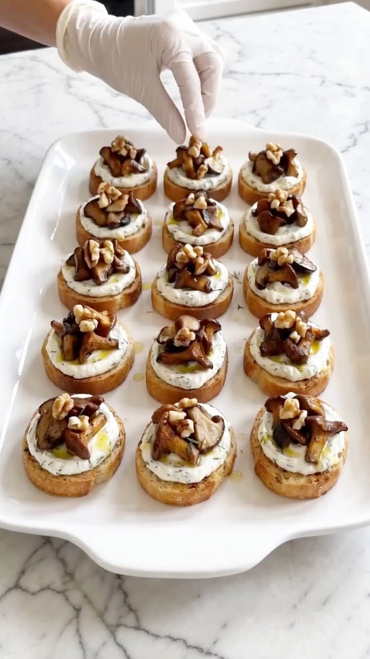 A gloved hand places mushroom and ricotta crostini onto a white serving platter.