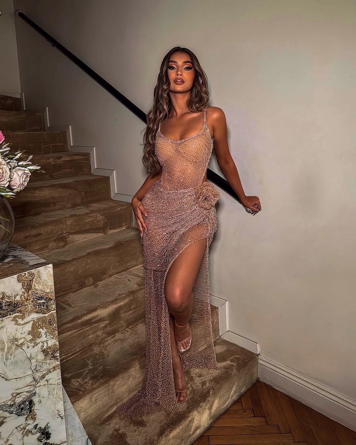 A woman in a sparkly, sheer, floor-length embellished dress posing on a marble staircase.