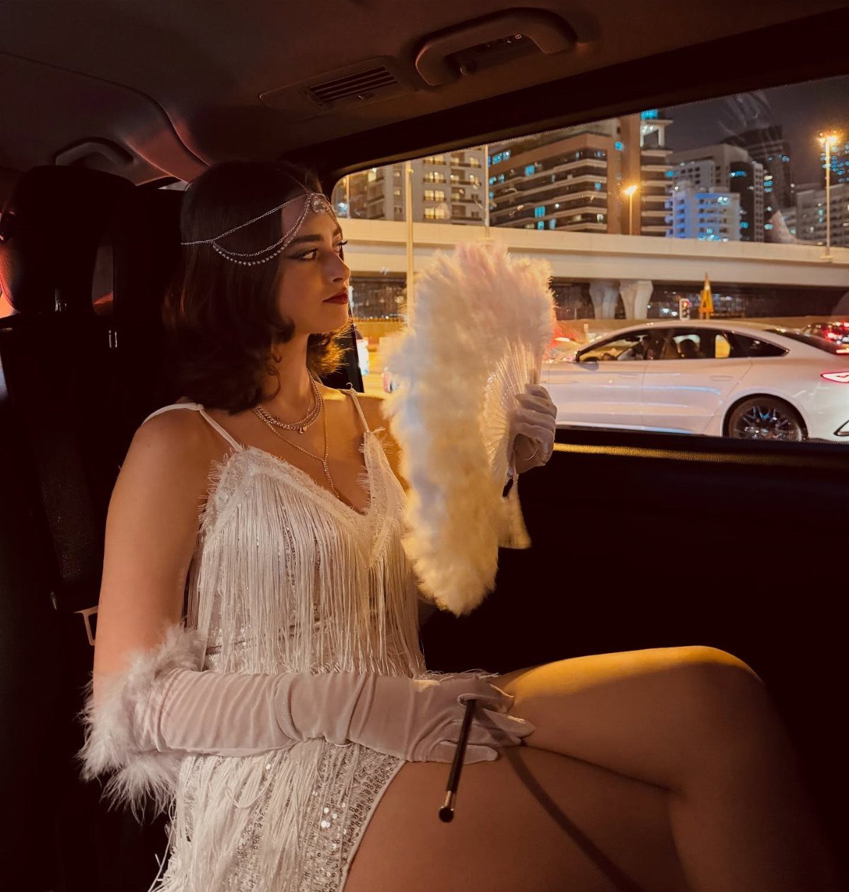 Woman in a white flapper dress wearing a headband and holding a feather fan.