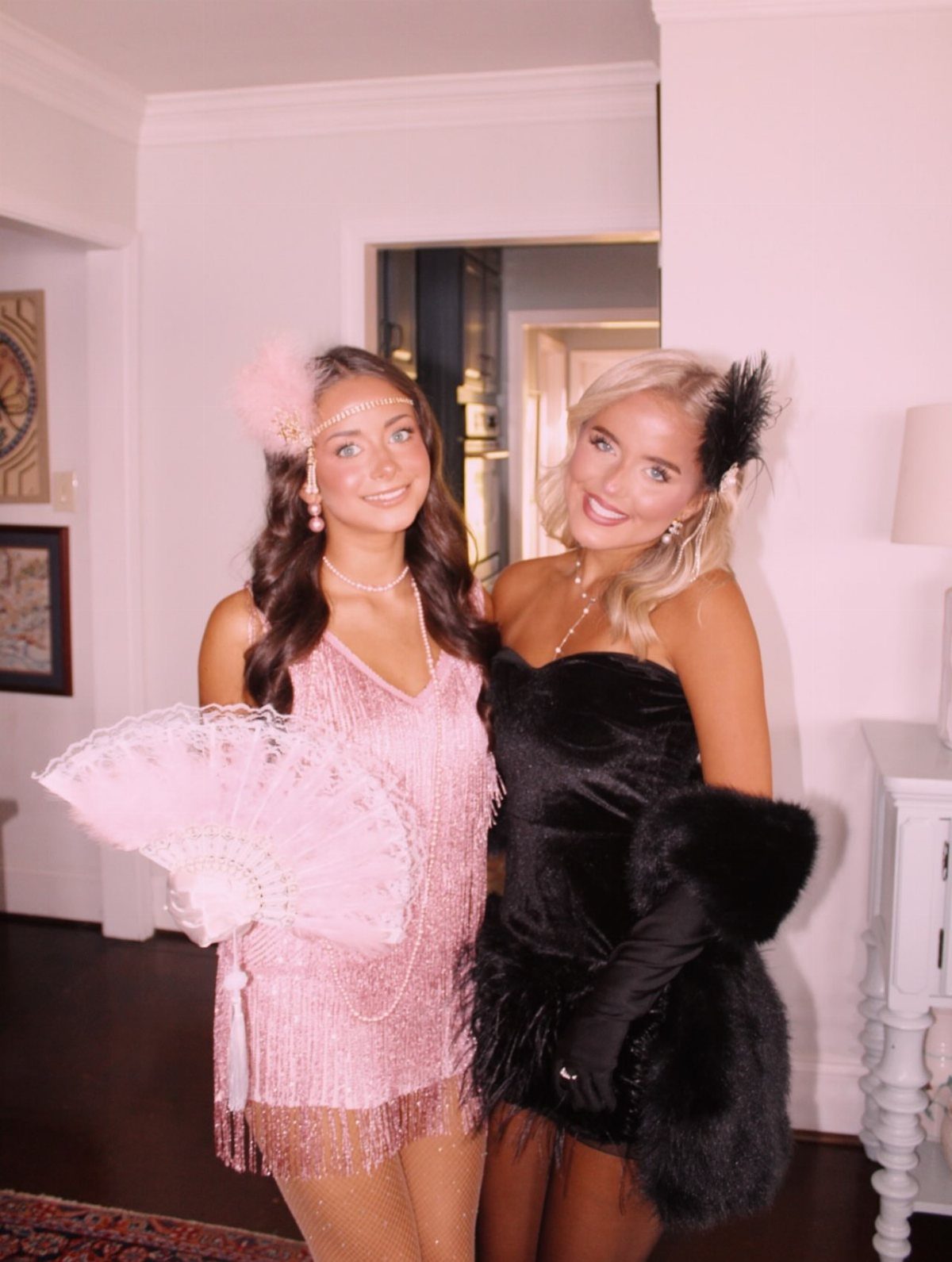 Two women smiling while posing in pink and black sequined flapper costumes with feather accessories.