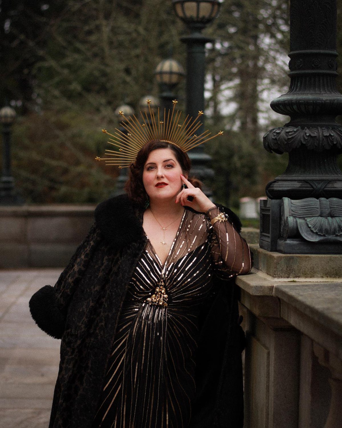 A woman in a black and gold sequined flapper dress and sunburst headpiece posing outdoors.